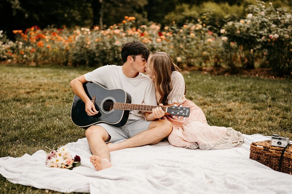 A young man and girl sitting on a white blanket in a garden, sharing a kiss while the man plays an acoustic guitar. Flowers and trees are in the background, with a basket and a camera beside them.