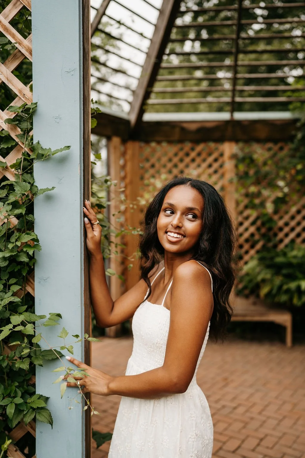 A young woman with long, wavy dark hair smiling and posing outdoors near a light blue pillar with green vines, in a garden setting with lattice and trees in the background.