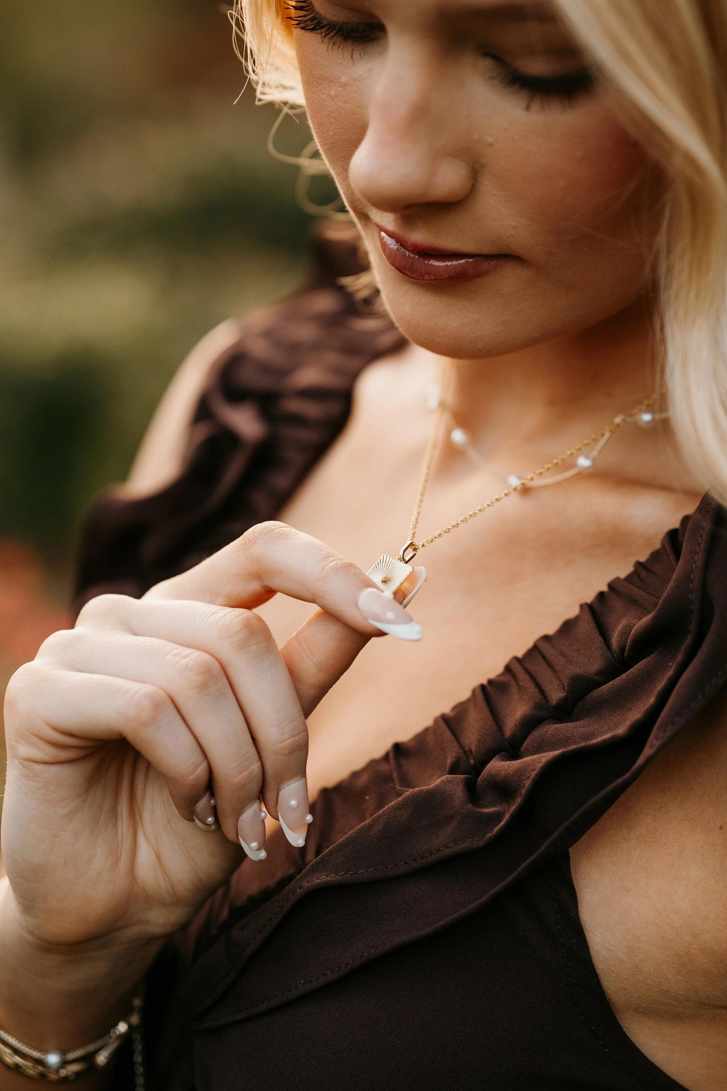 A woman with blonde hair wearing a black outfit with ruffled details, holding a gold necklace with a pendant, her nails painted white with pearl accents, outdoors during golden hour.