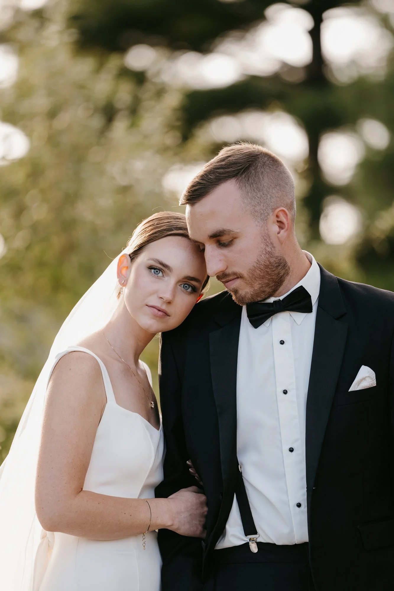 A bride and groom embrace outdoors during their wedding, with the bride wearing a white dress and veil, and the groom in a tuxedo with a bow tie, both looking serene and close together.