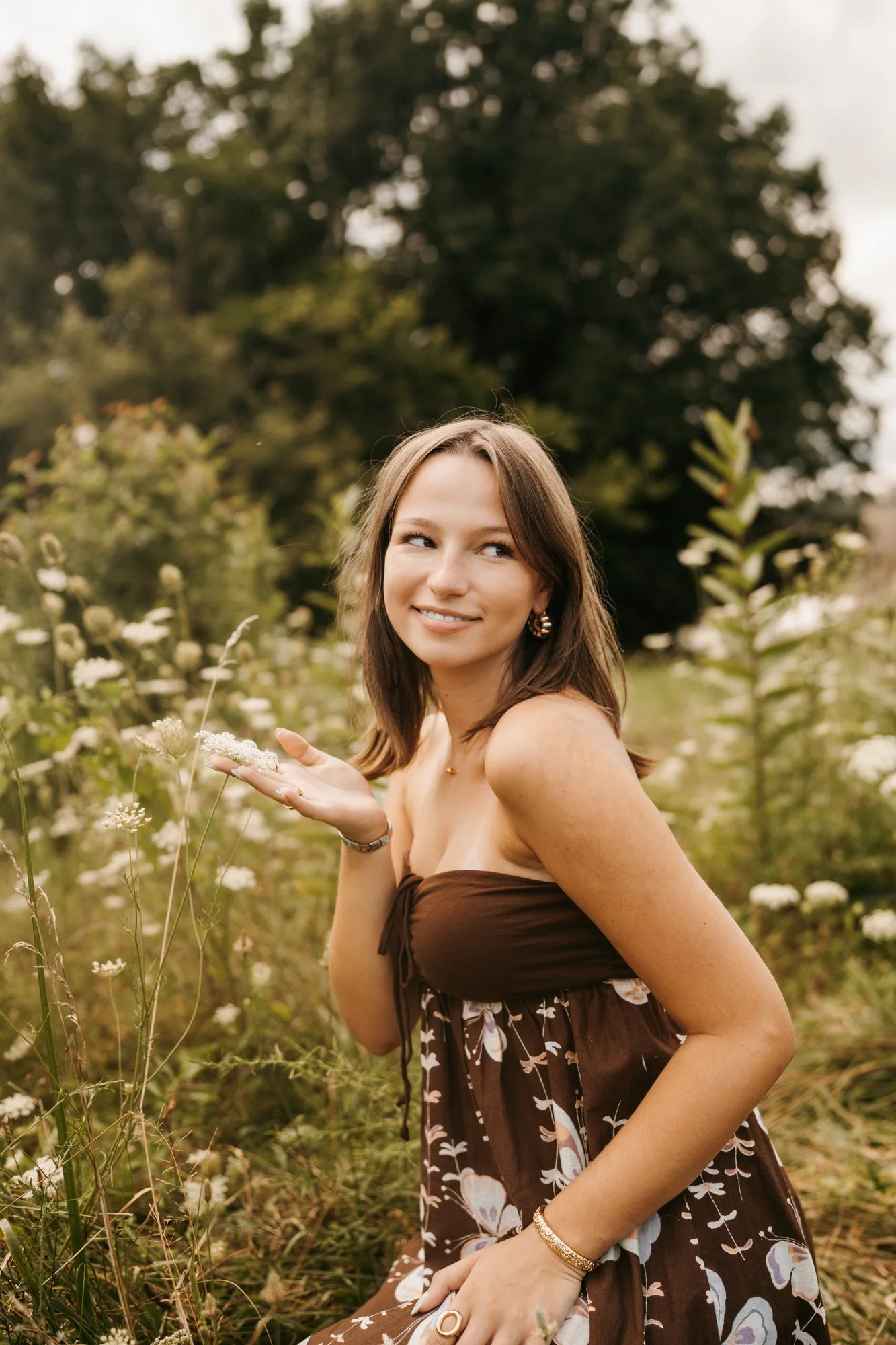 A woman in a brown floral dress sitting in a field of white wildflowers, holding a flower in her hand and smiling at the camera.