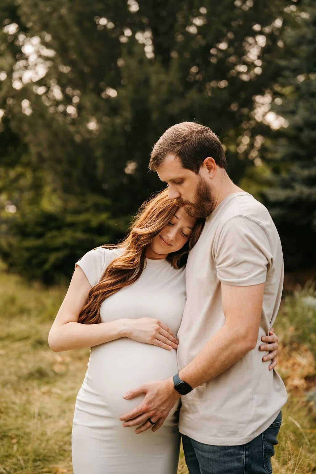 A pregnant woman and a man embracing outdoors in a park or garden, with trees in the background, during sunset or late afternoon.