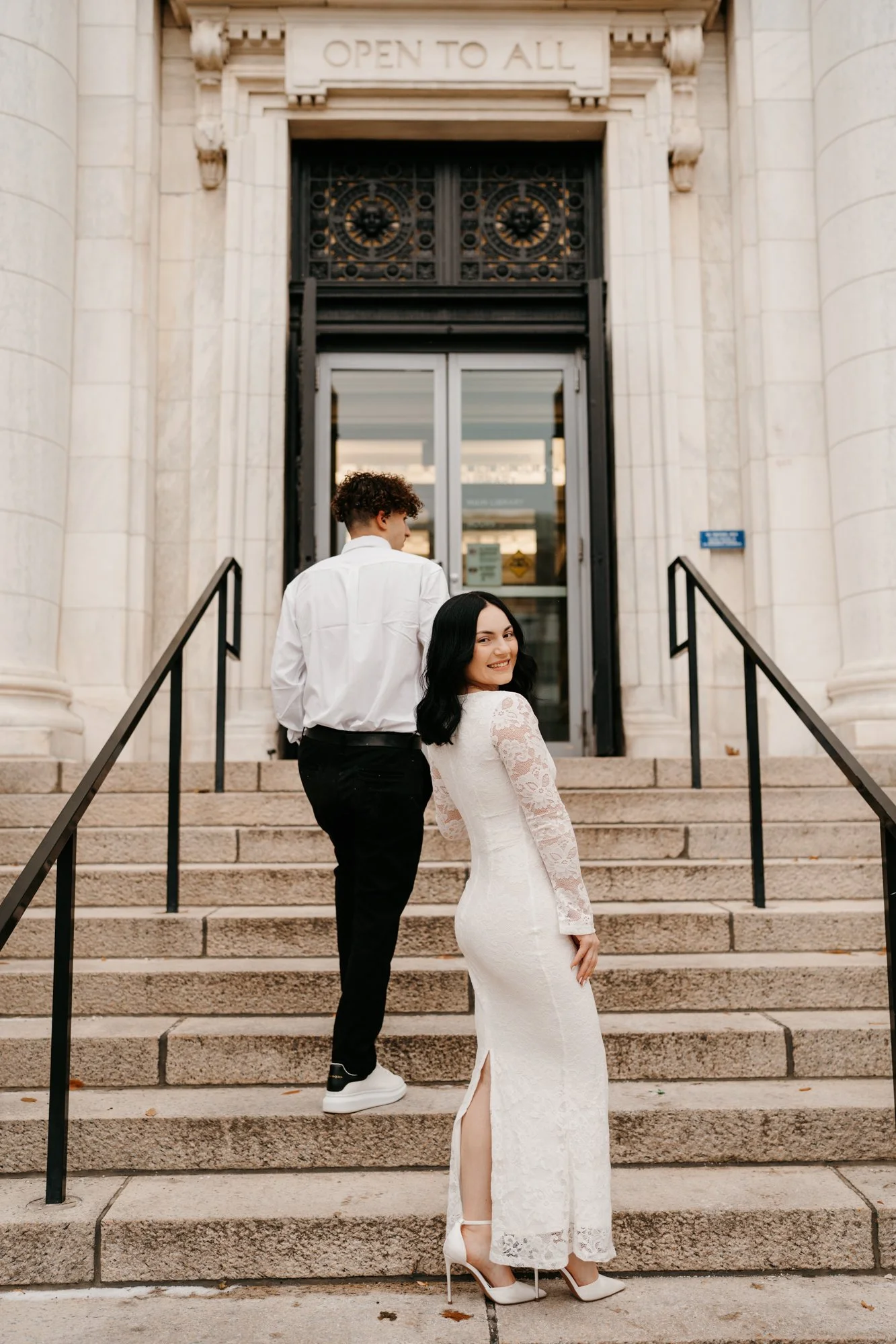 A woman in a white lace dress and heels smiling at the camera as she stands on the steps of a building with a man in black pants and white shirt walking away.