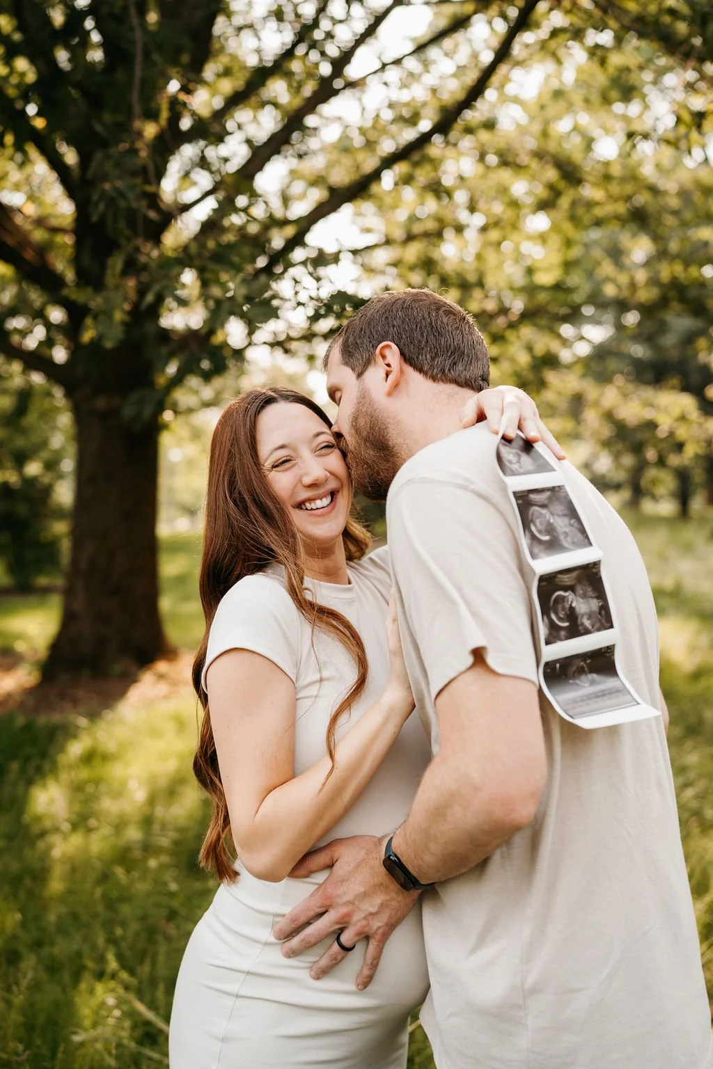 A couple outdoors, the woman is smiling as the man kisses her forehead, with ultrasound images hanging from his neck. The scene is set in a park with grass and a large tree in the background, during golden hour.