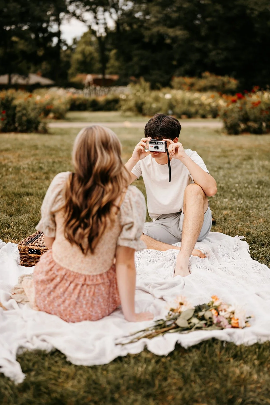 A boy takes a photo of a girl with a vintage camera while sitting on a blanket in a park with flowers and trees.