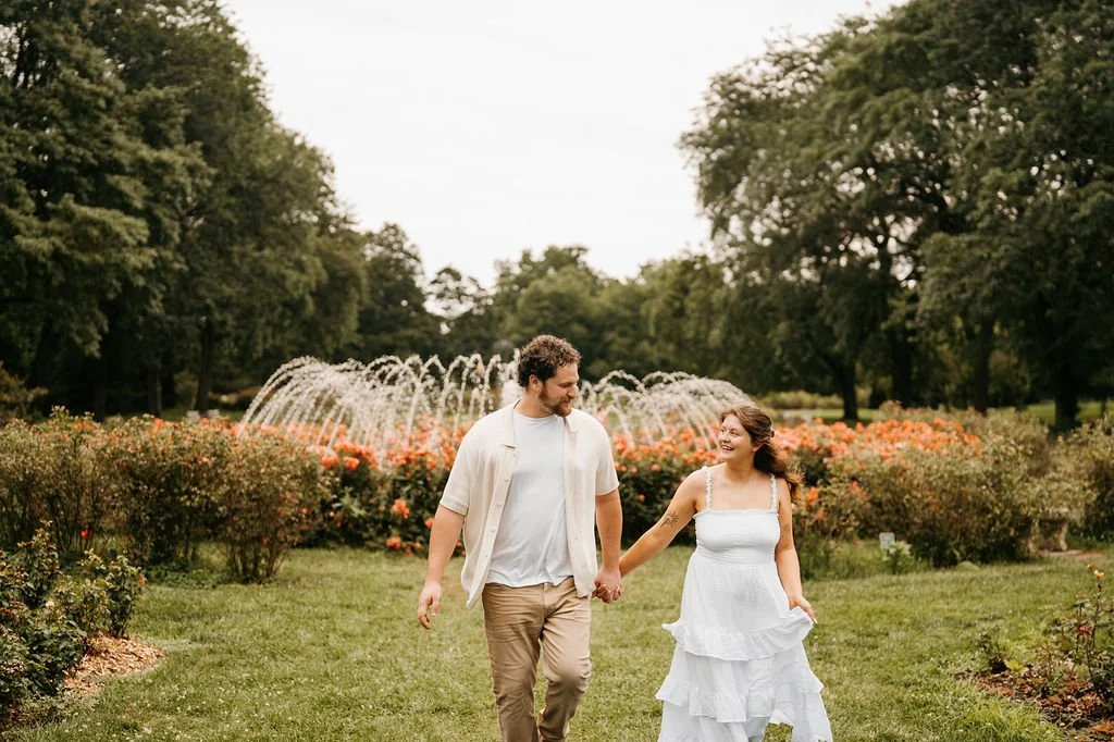 A couple walking hand in hand in a garden with a water fountain and blooming flowers.