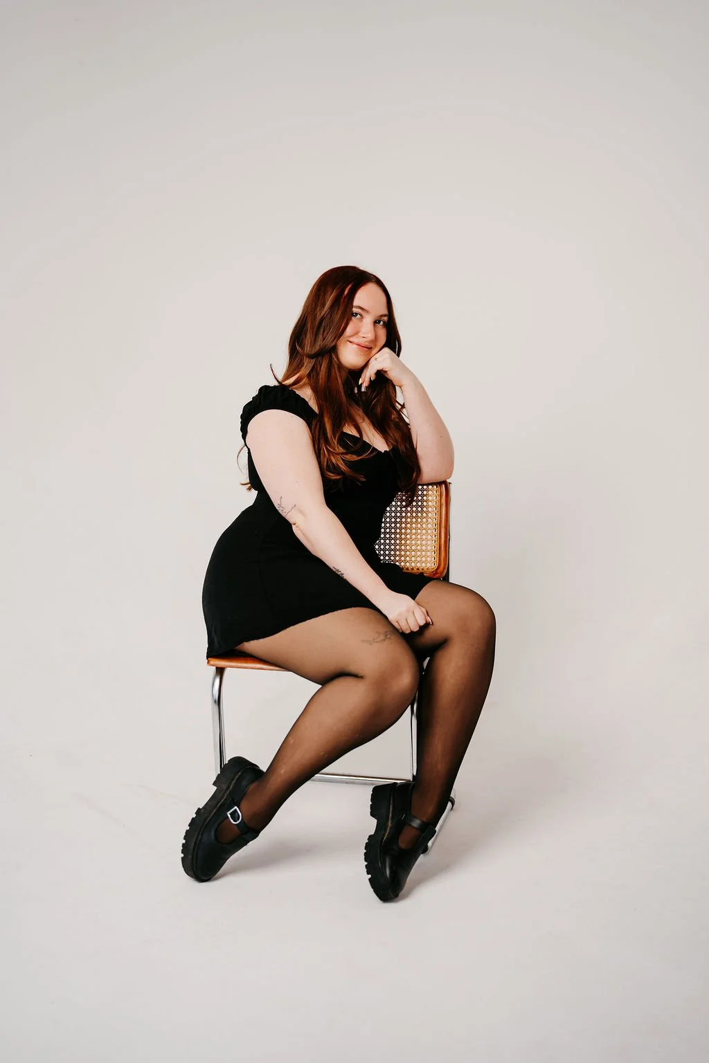 A woman with long, wavy brown hair wearing a black dress, black tights, and chunky black shoes, sitting on a chair against a plain white background, smiling and resting her chin on her hand.