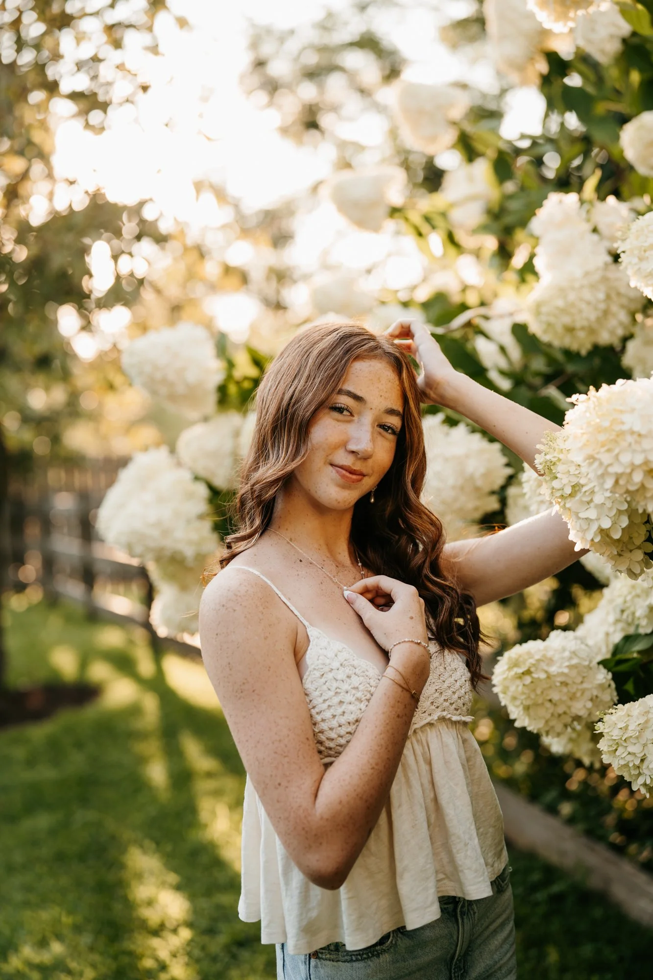 A young woman with long wavy brown hair and freckles stands in a garden with white hydrangea flowers, holding her hair with one hand and touching her necklace with the other, during golden hour.