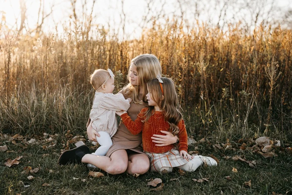 A woman and two young girls sit on the ground in a field of fallen leaves during autumn, with trees in the background. The woman holds the younger girl, and the older girl sits beside them, all dressed in warm clothing.