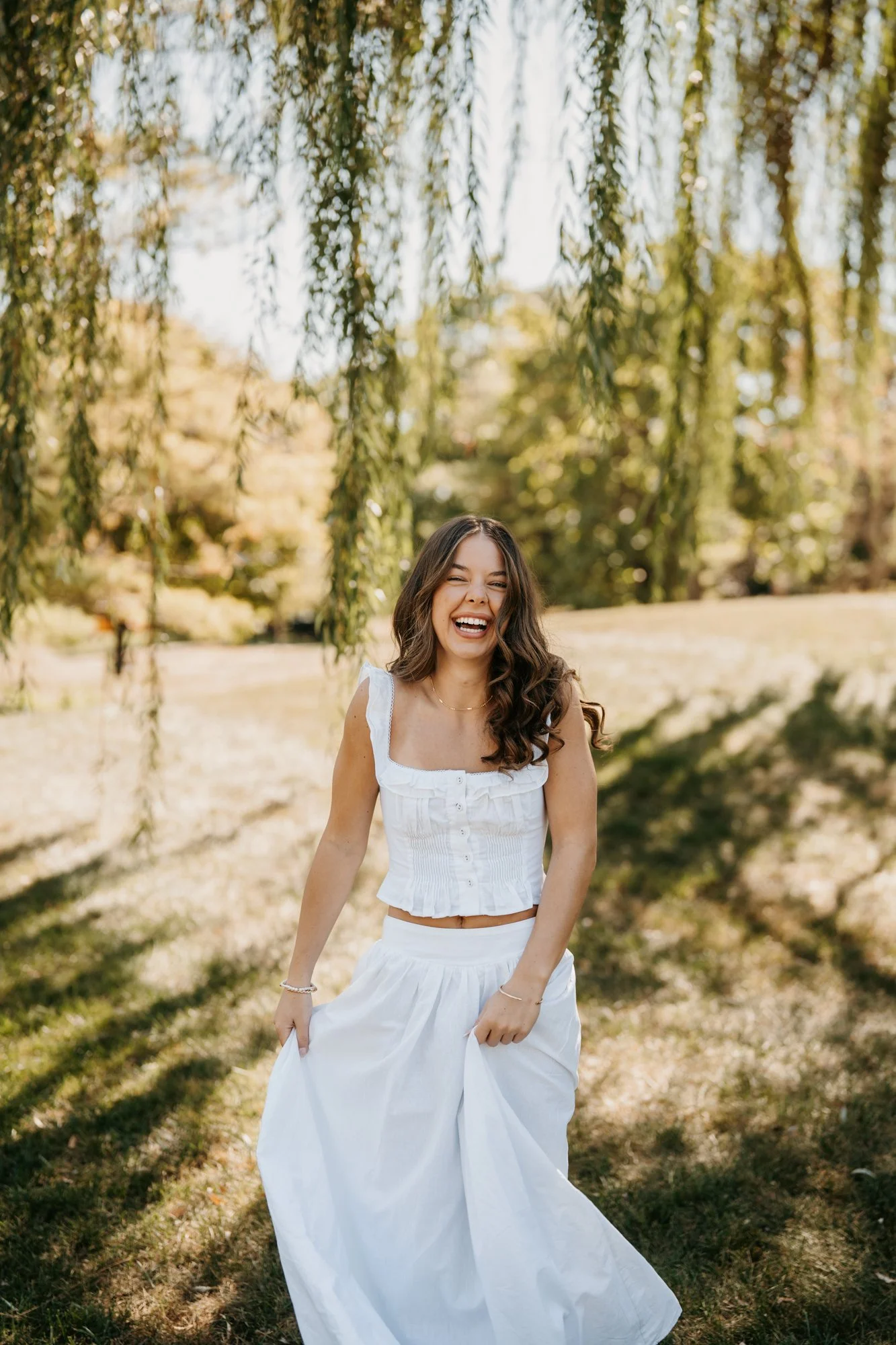 Woman in white dress laughing outdoors under a tree in sunlight.