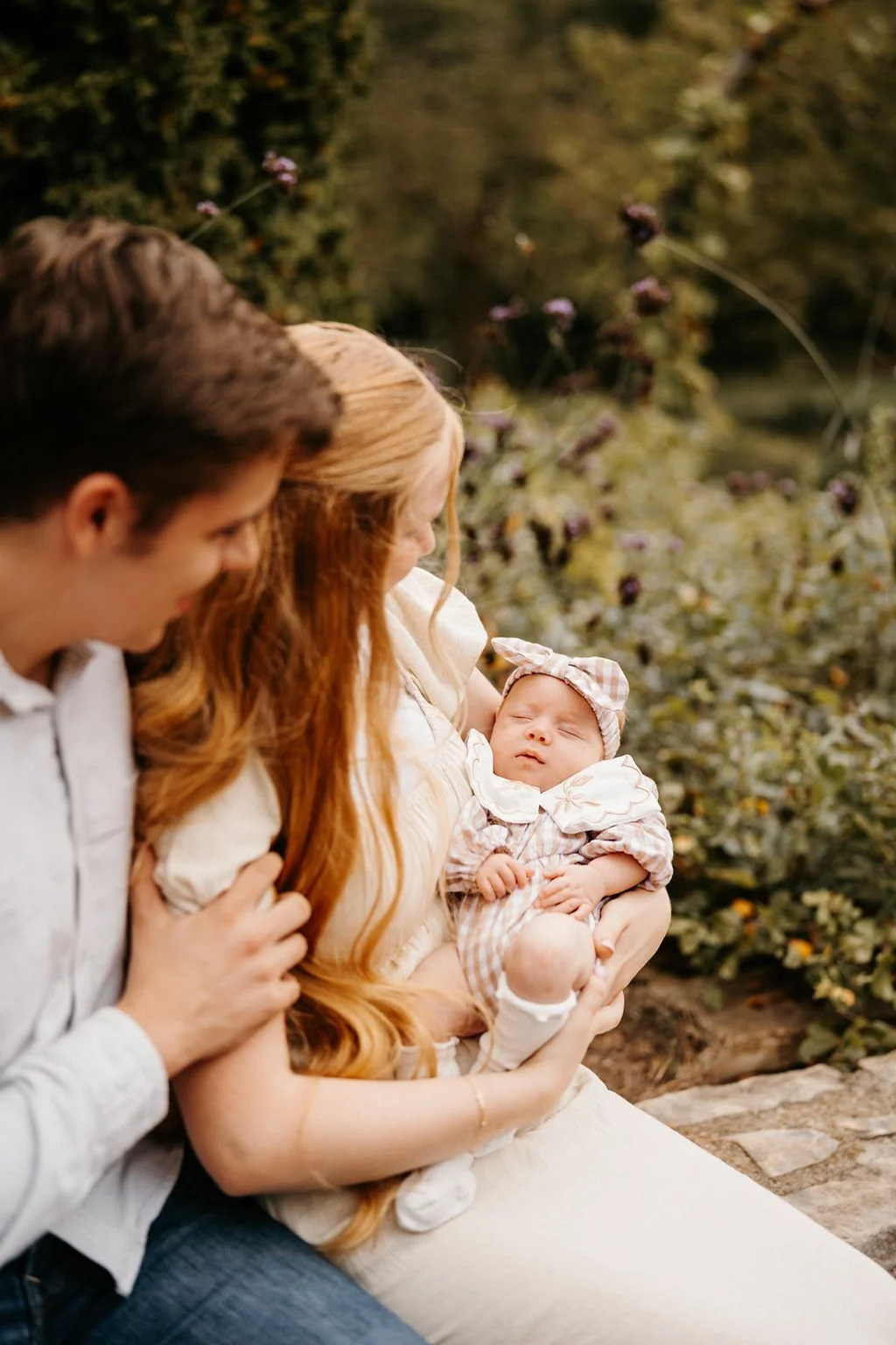 A woman holding a sleeping baby girl outside, with a man nearby looking at the baby.