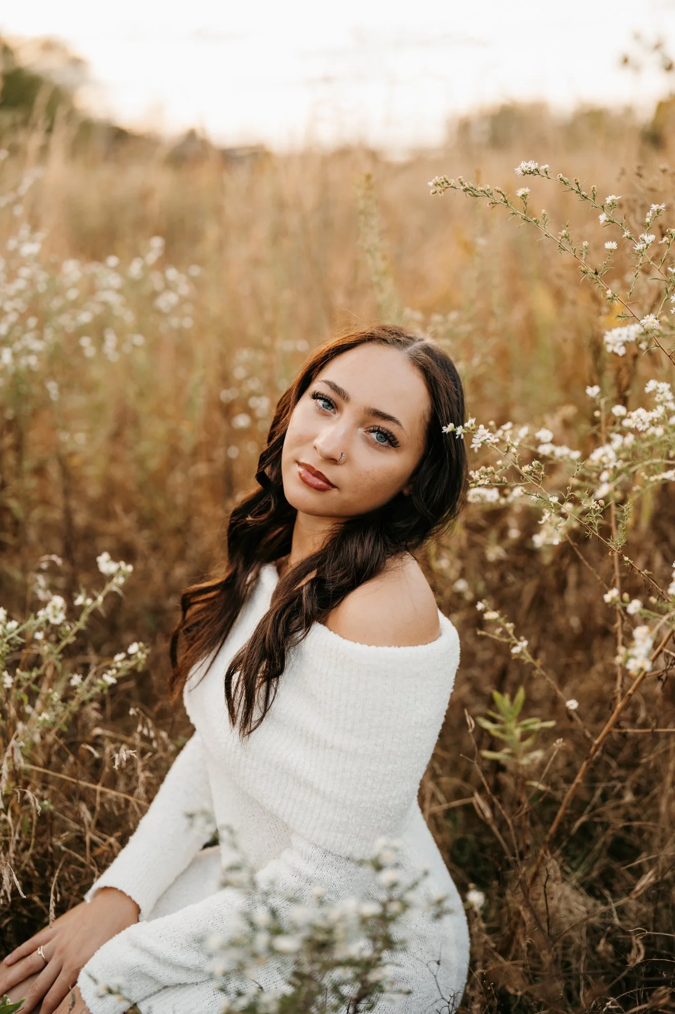 A young woman with long dark hair and blue eyes sitting in a field of tall grasses and white wildflowers during sunset, wearing a white off-the-shoulder sweater.
