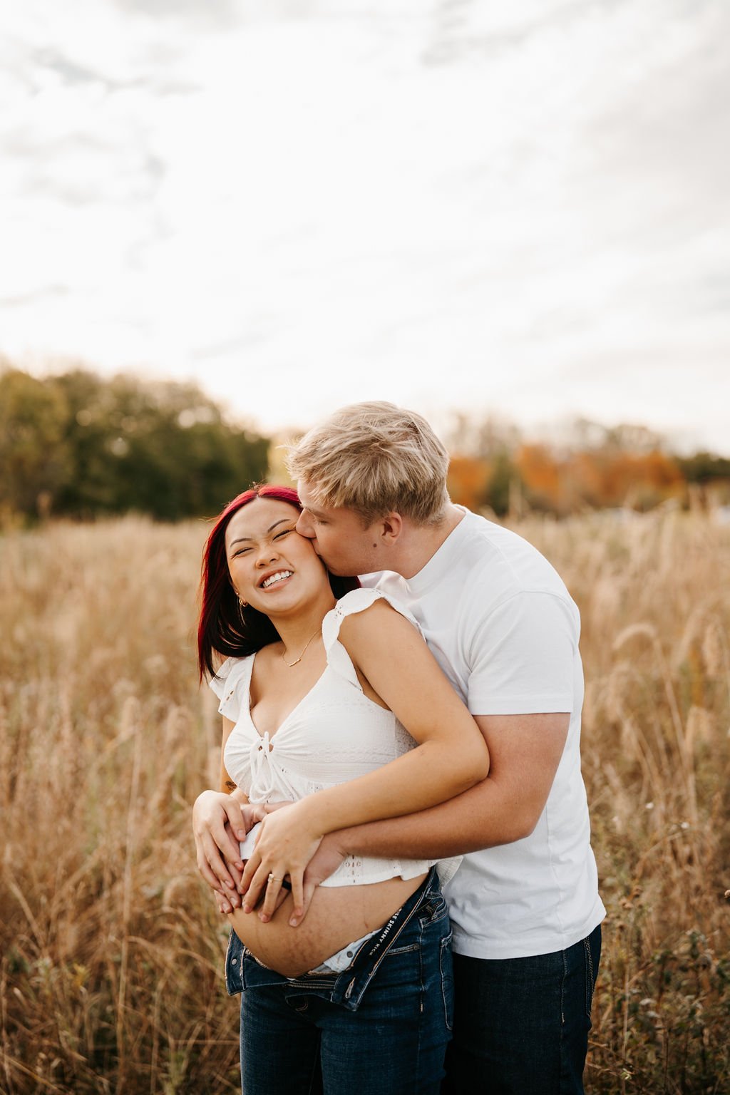 A pregnant woman with red hair and a white top stands outdoors in a field, smiling while a man with blond hair kisses her cheek.