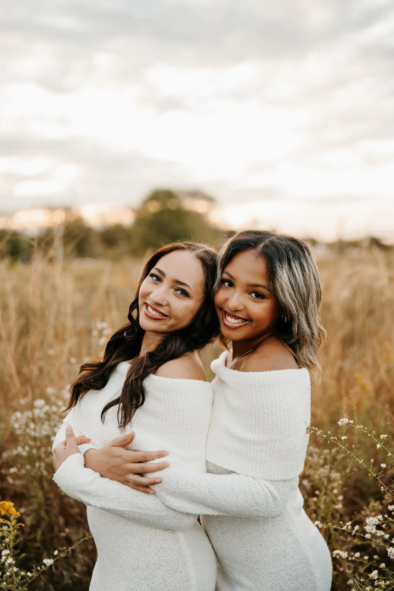 Two women with dark and light skin tones embracing outdoors in a field of tall grass and wildflowers at sunset, wearing matching white off-the-shoulder sweaters, smiling and looking at the camera.