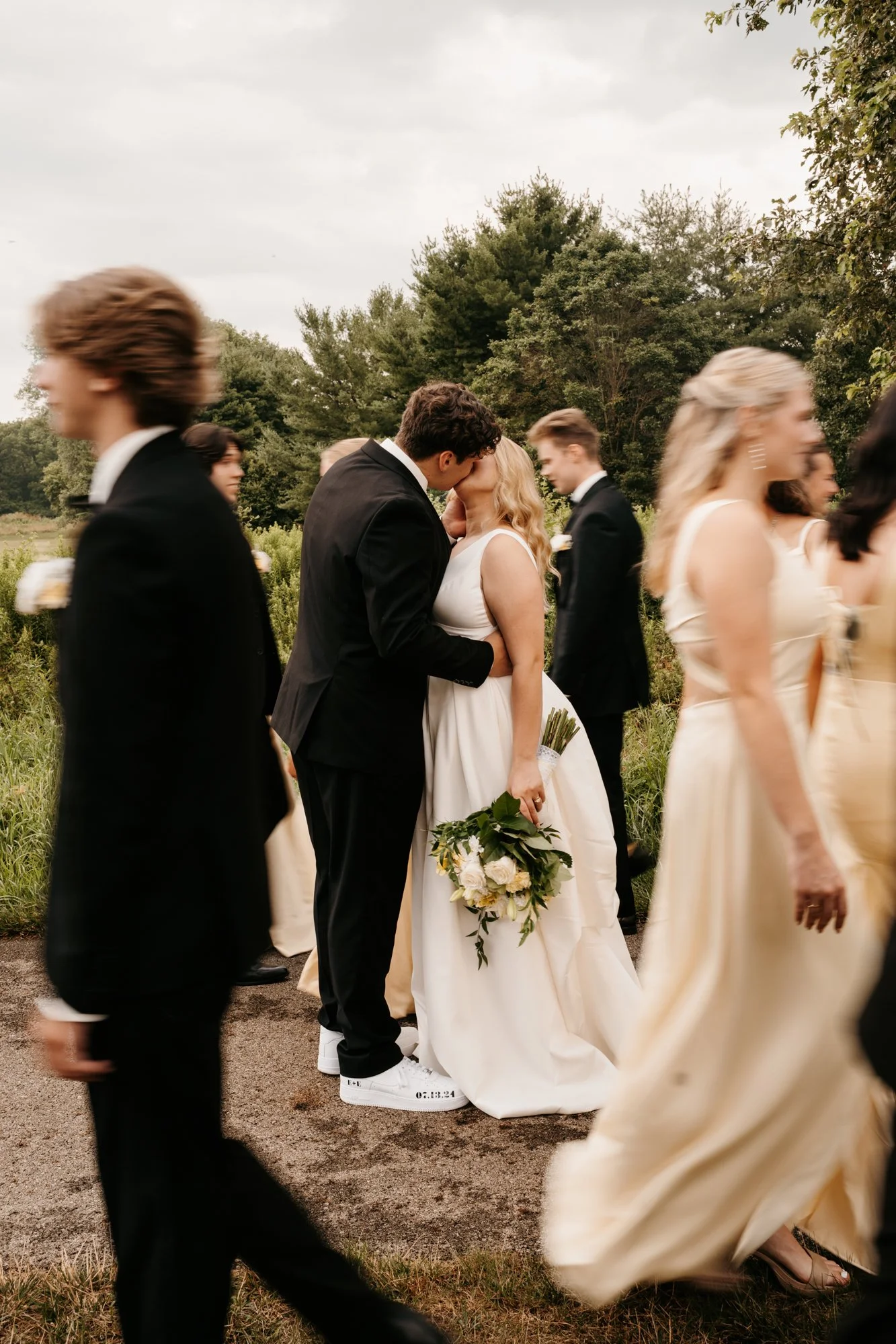 Bride and groom sharing a kiss during their wedding, surrounded by wedding guests walking outdoors.
