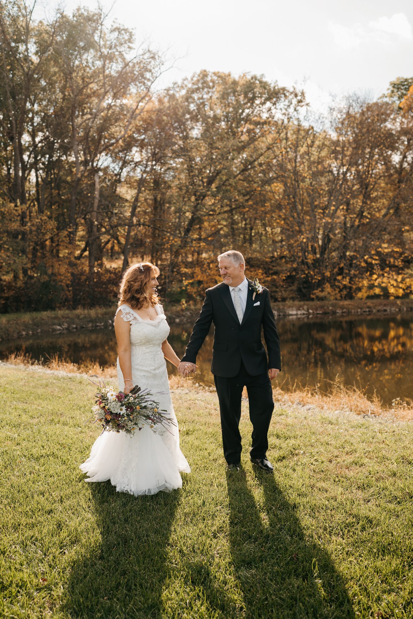 A bride and groom holding hands and walking outdoors near a pond, during a sunset with fall foliage in the background.