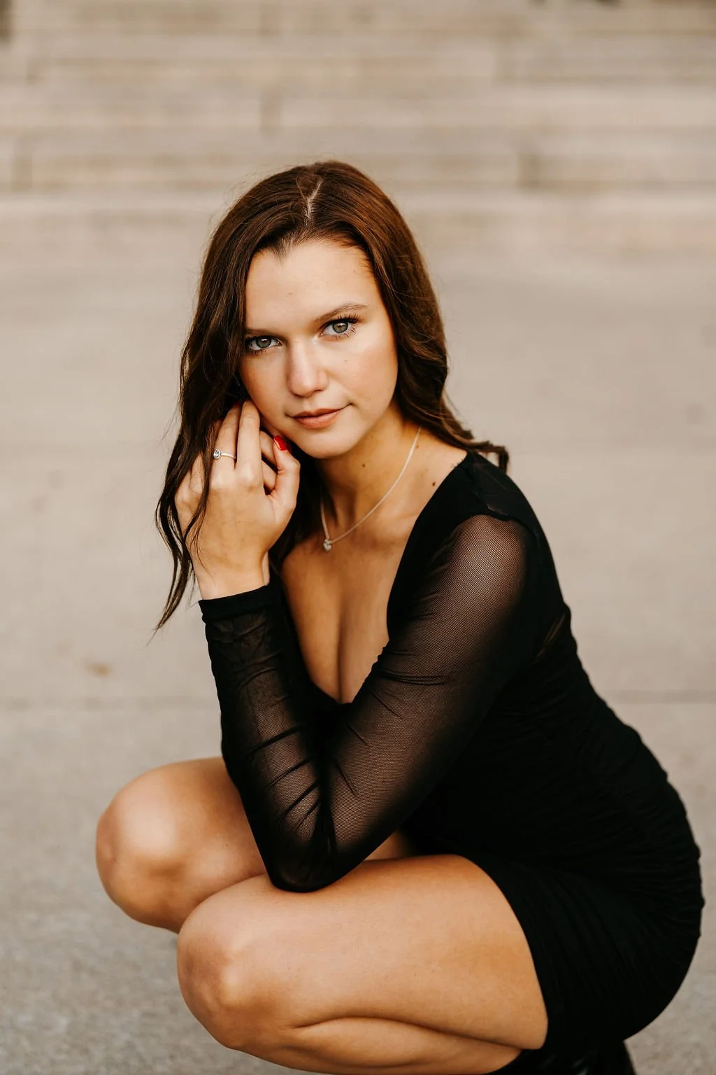 Portrait of a woman with shoulder-length brown hair wearing a black dress with sheer sleeves, sitting in a squatting pose with one hand near her face, against a neutral background.
