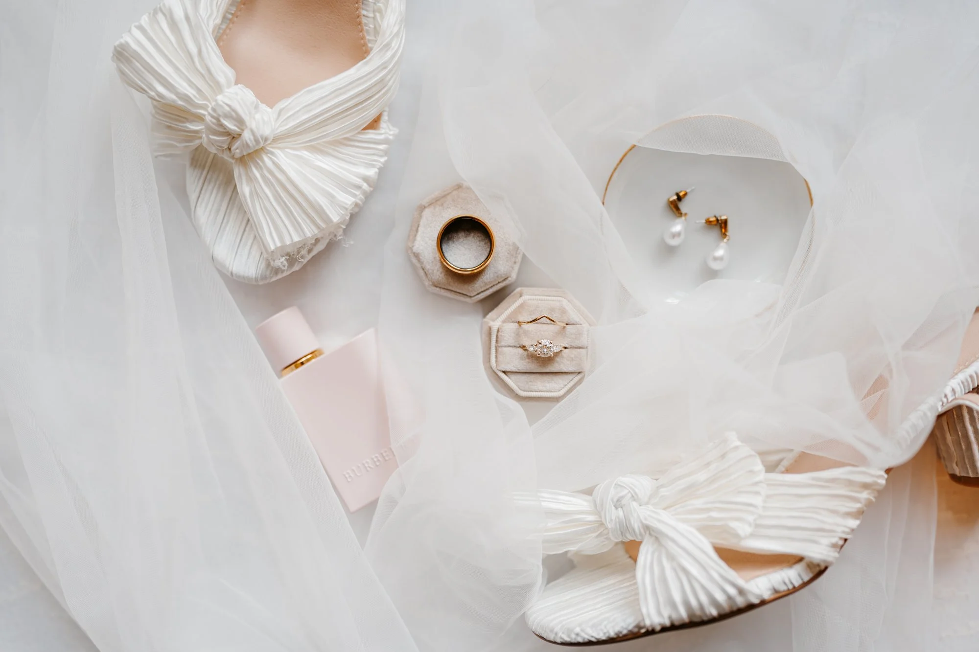 Flat lay of jewelry and accessories including rings, earrings, perfume, and shoes with white fabric and tulle.