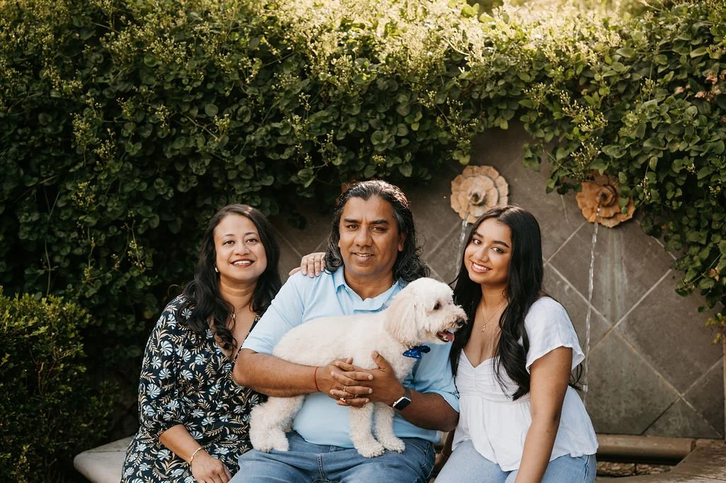 A family of three adults and a dog posing for a photo outdoors, with lush green foliage and decorative flowers in the background.
