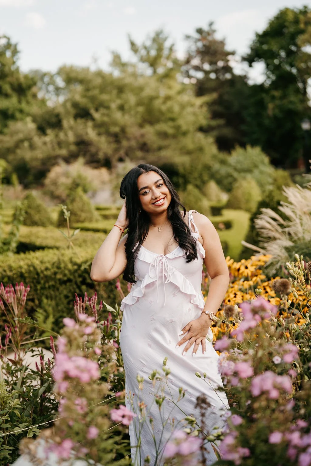 A young woman with dark hair smiling outdoors among colorful flowers and greenery.