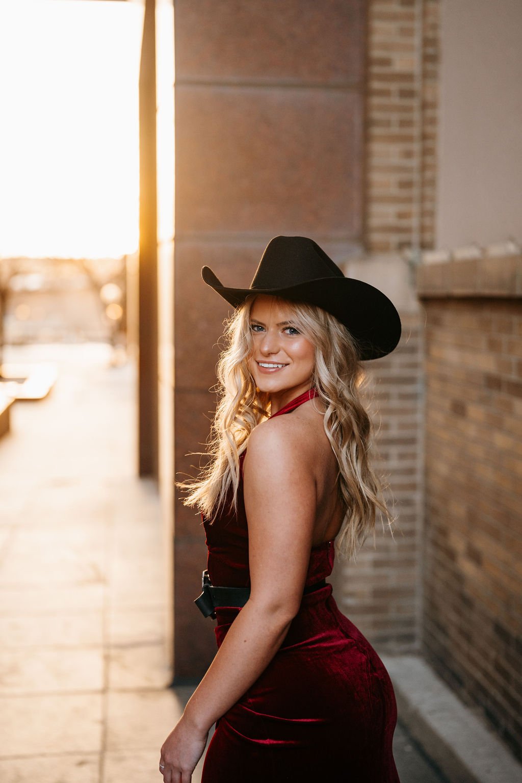 A woman with long blonde hair smiling, wearing a black cowboy hat and a red dress, standing in an alleyway with brick walls at sunset.