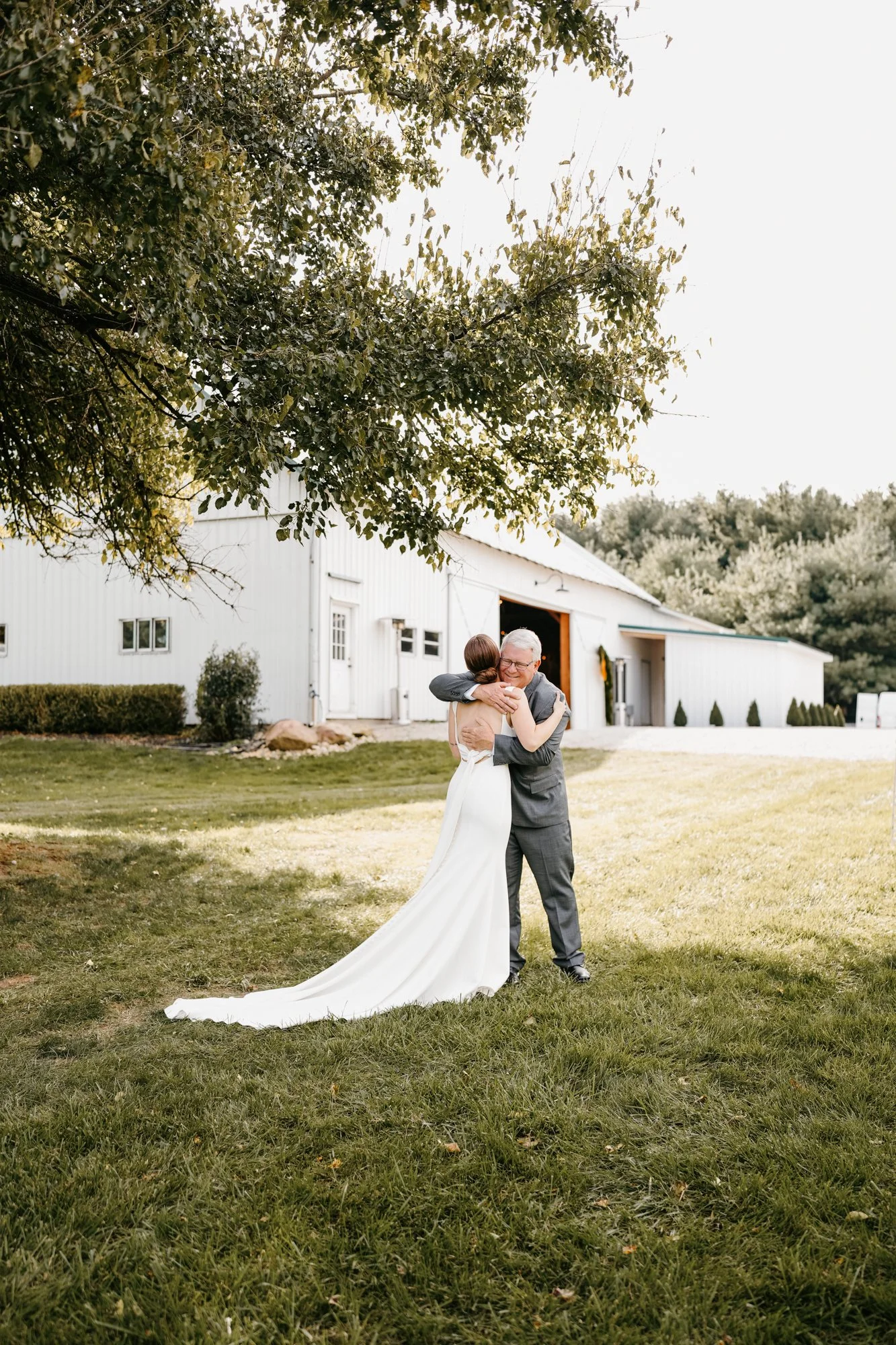 A bride and an older man embracing outdoors in a grassy area near a white barn, with trees in the background.