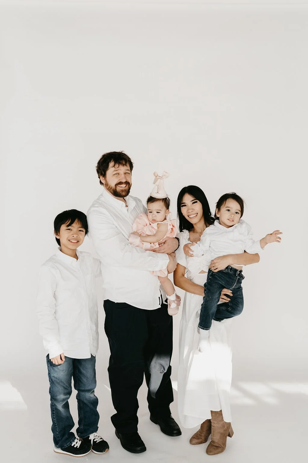 A diverse family of five standing against a plain white background, including a man, woman, two young girls, and a boy, all smiling and dressed in white shirts and casual clothes.