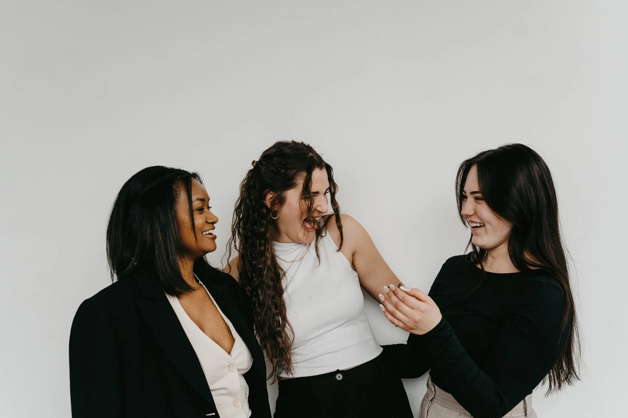 Three women laughing and smiling together against a plain white background. The woman on the left has dark skin and black hair, wearing a black blazer and white blouse. The woman in the center has light skin, long curly brown hair, wearing a white sl