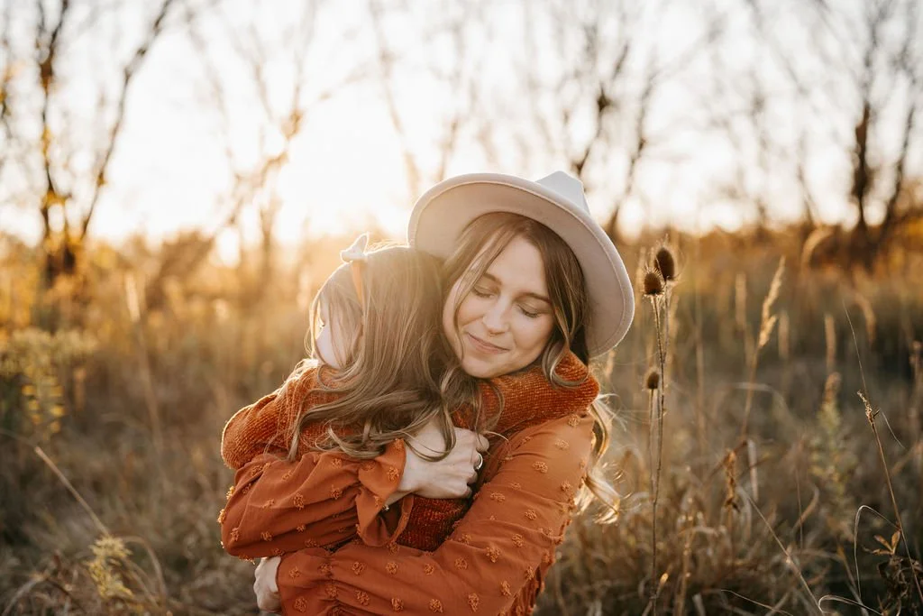 A woman wearing a wide-brimmed hat hugs a young girl in an outdoor field at sunset, surrounded by tall dry grass and trees.