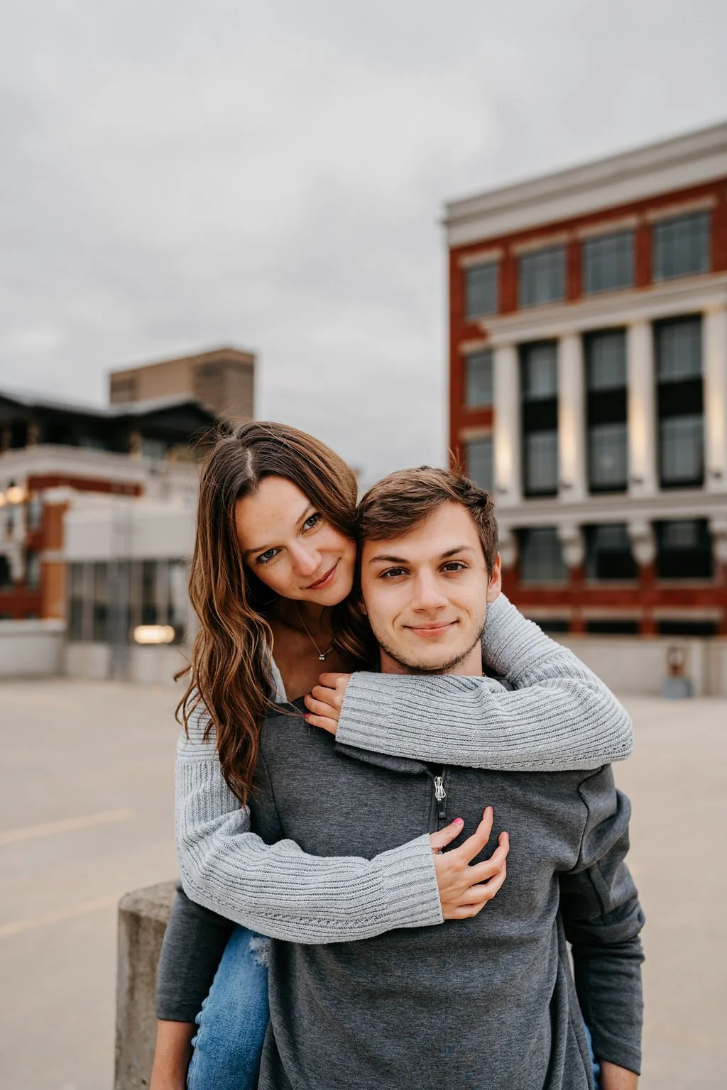 A young woman with long brown hair embracing a young man with short brown hair, both smiling, standing outdoors in an urban area with brick and glass buildings in the background.