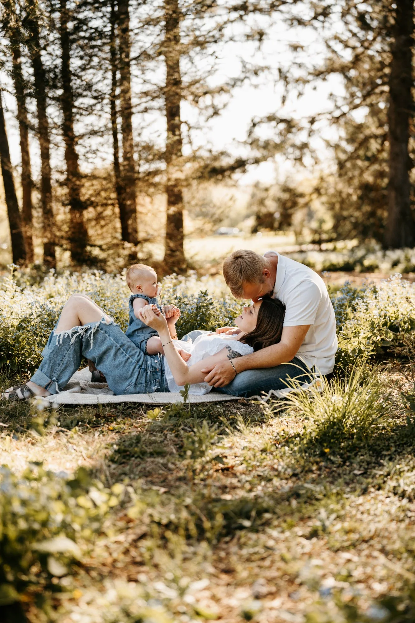 Family lying on blanket in a forest, with mother, father, and two children, enjoying a sunny day.