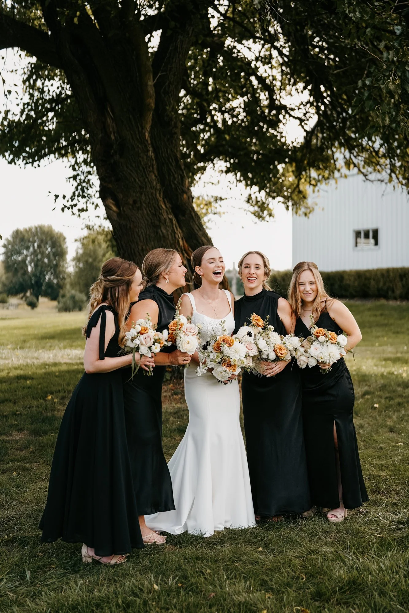 A bride and four bridesmaids stand outdoors under a large tree, all holding bouquets of flowers, laughing and smiling during a wedding celebration.
