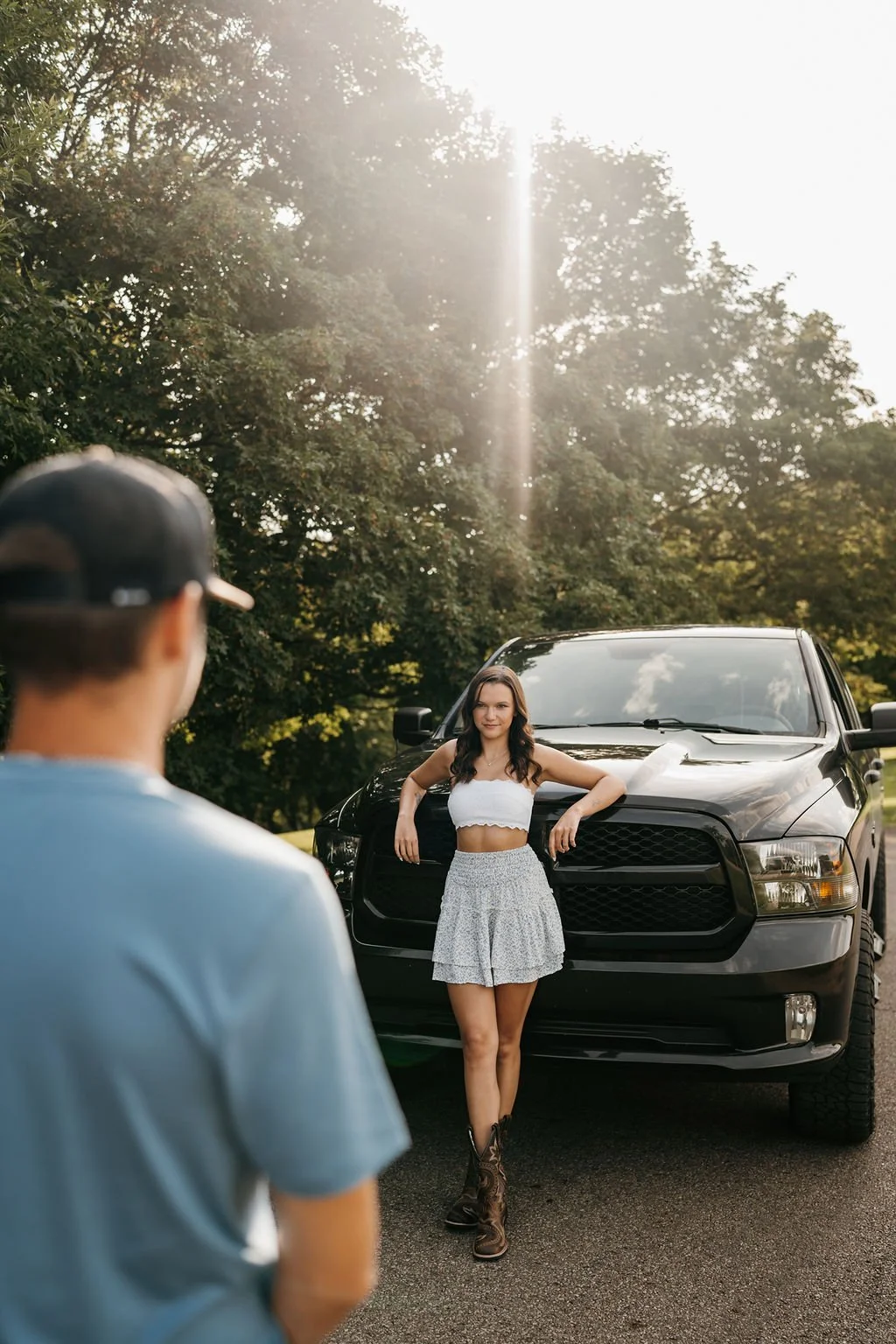 A woman in a white crop top and gray skirt posing in front of a black truck on a sunny day with trees in the background. A person in the foreground is looking at her.
