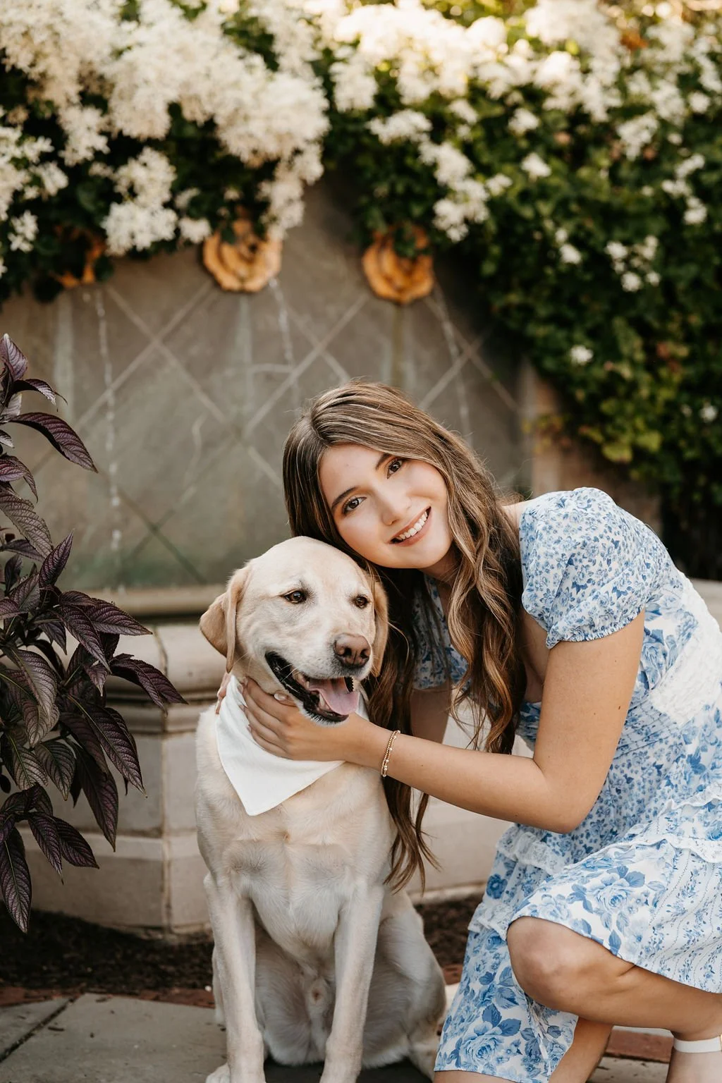 A young woman with long brown hair and a blue and white dress is hugging a yellow Labrador Retriever dog with a white bandana around its neck. They are outdoors near a flower garden with white flowers and greenery.