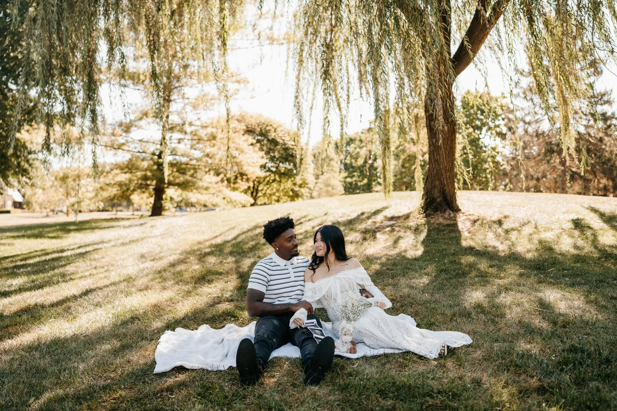 Couple sitting on a white blanket under a large tree in a park on a sunny day.