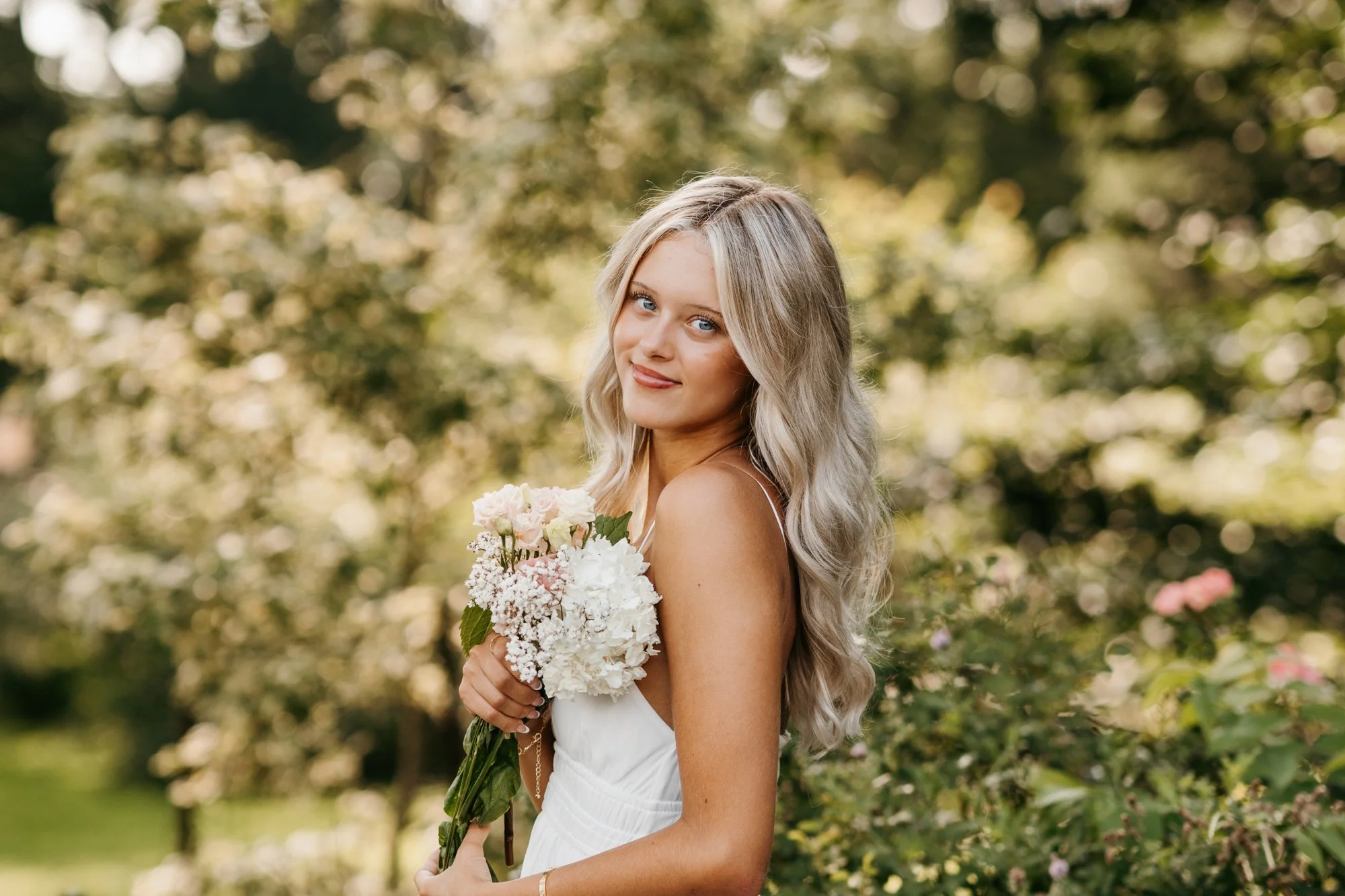 A young woman in a white dress holding a bouquet of flowers outdoors with trees and greenery in the background.