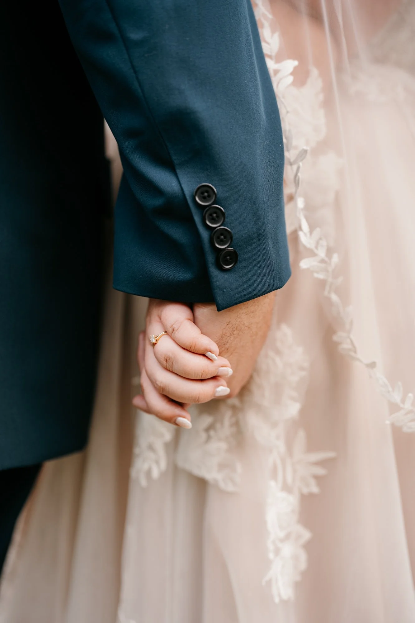 Close-up of a bride and groom holding hands, with the groom dressed in a navy suit and the bride wearing a white gown with floral lace detail.