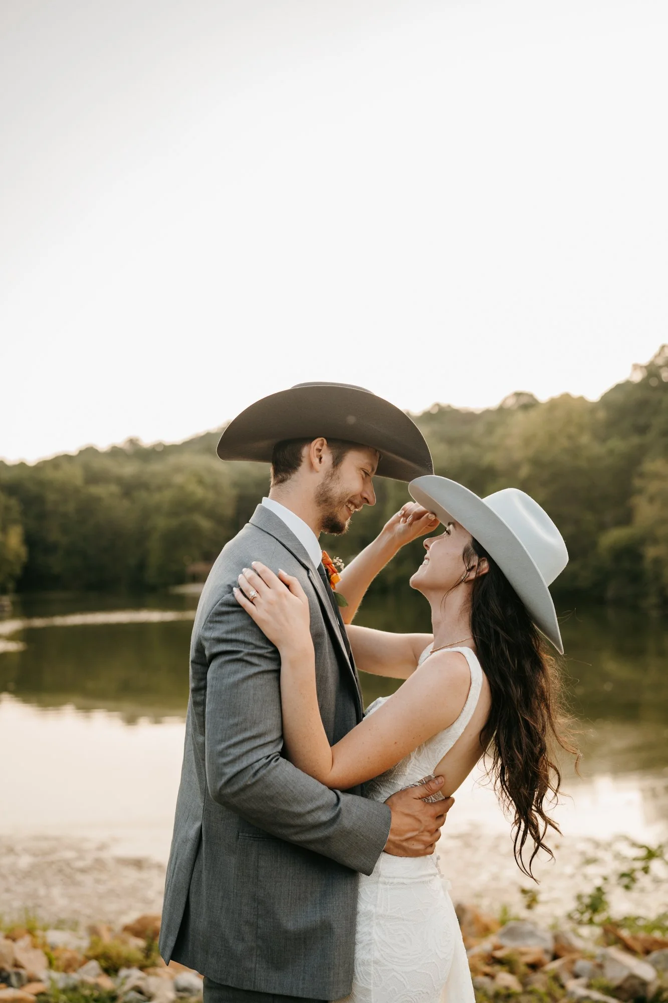 A couple in wedding attire embracing outdoors near a lake, both wearing cowboy hats, smiling at each other.