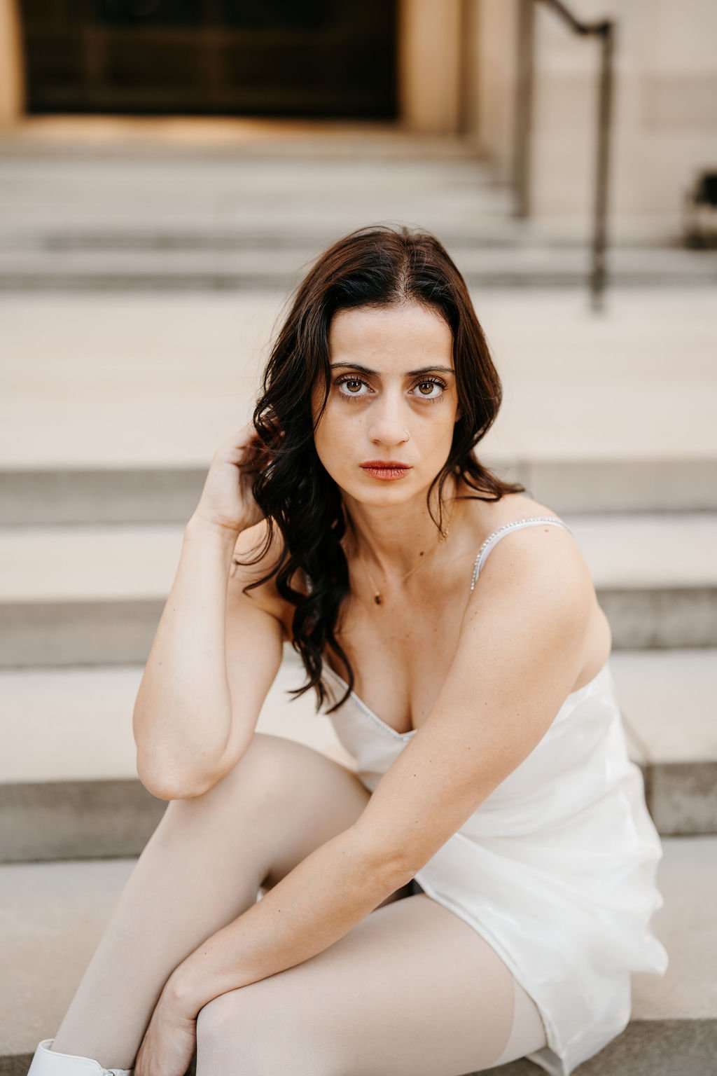 A young woman with long dark hair and light skin, sitting on outdoor steps, wearing a white satin dress and beige tights, looking directly at the camera.