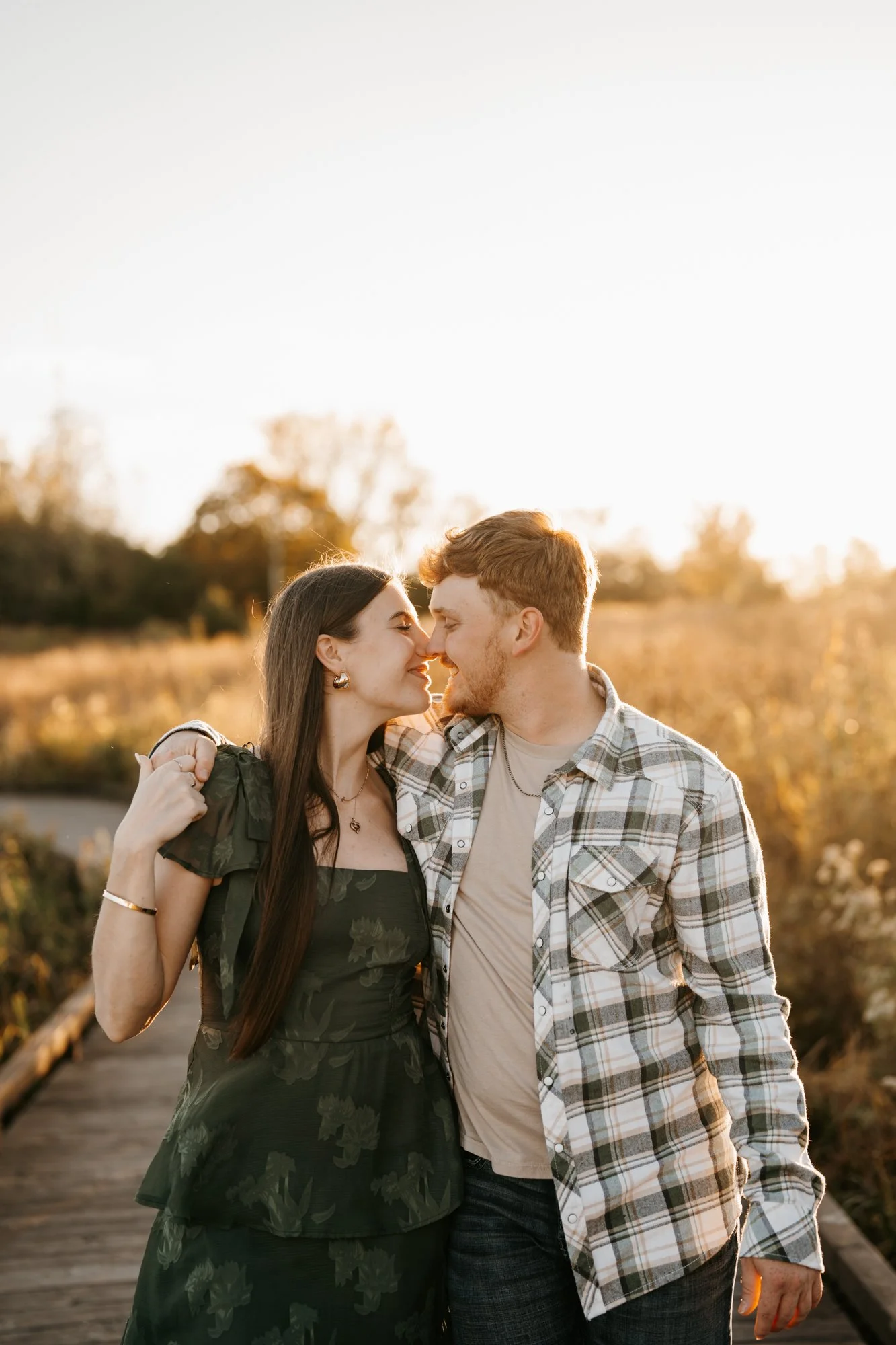 A young couple standing close, holding each other with their noses touching, smiling outdoors during sunset in a nature setting.