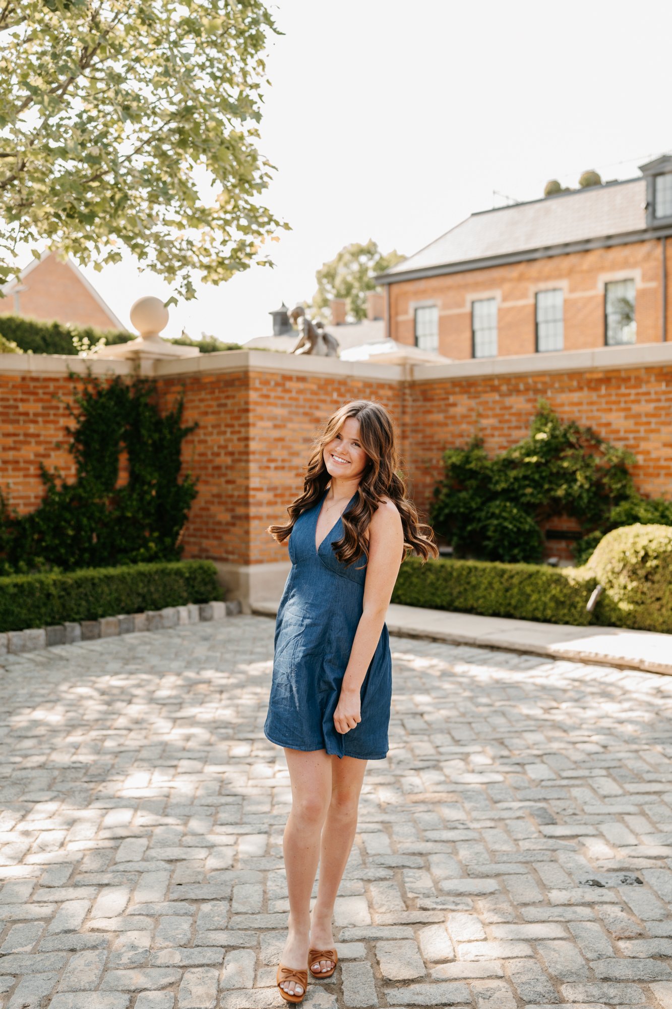 A young woman with long wavy brown hair standing on a cobblestone pathway, smiling, wearing a sleeveless blue dress and tan sandals. She is outdoors in a garden area with brick walls, green bushes, and trees in the background.