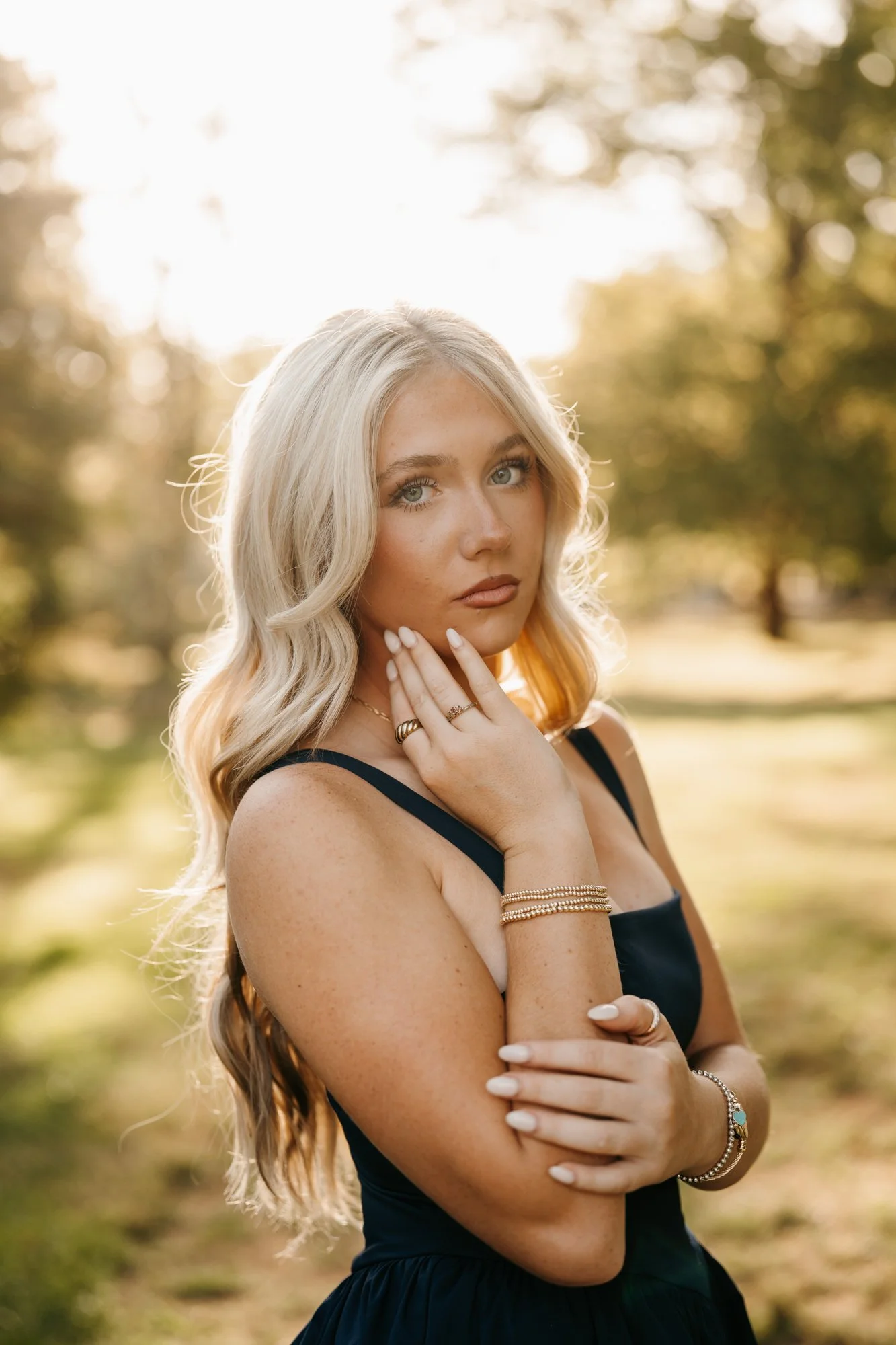 A young woman with long blonde wavy hair poses outdoors during golden hour with a backdrop of blurred trees and sunlight.