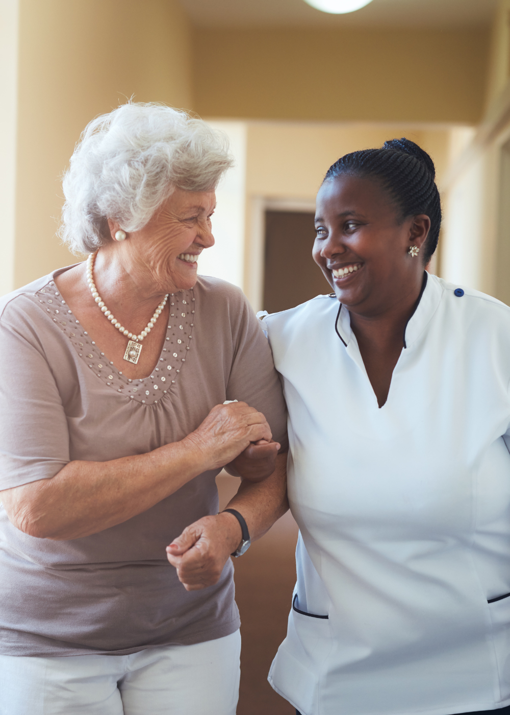 An elderly woman and a female healthcare worker smiling at each other in a care facility, holding hands.
