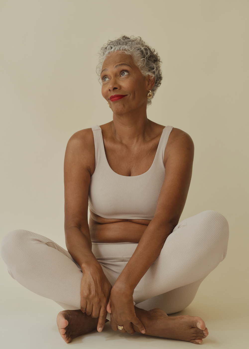 Older woman with gray curly hair sitting cross-legged on the floor, wearing a light beige sports bra and matching leggings, looking up with a playful expression.