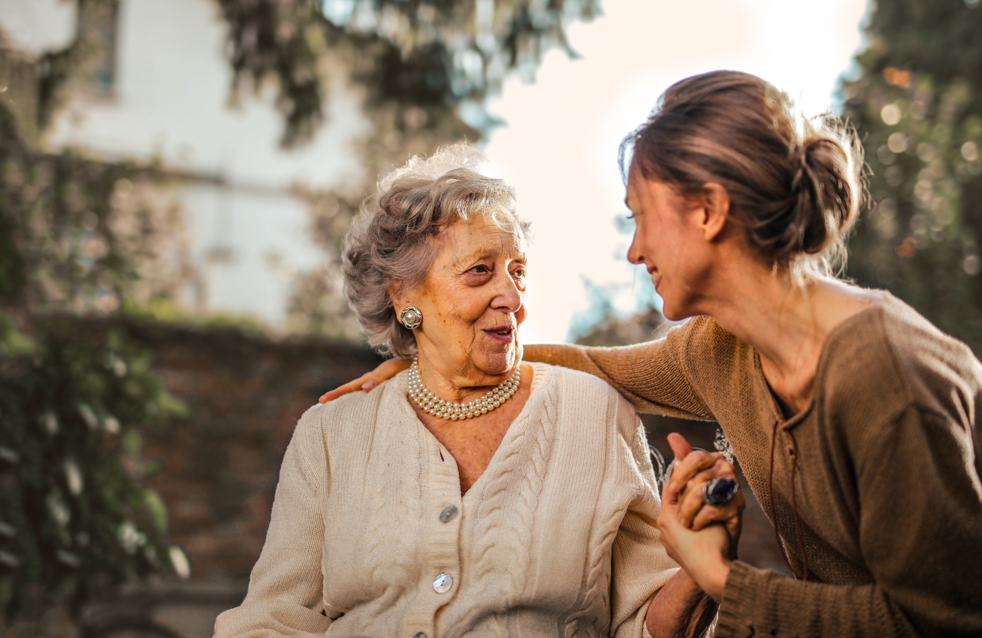 An elderly woman and a younger woman sharing a caring moment outdoors, smiling at each other, with trees in the background.