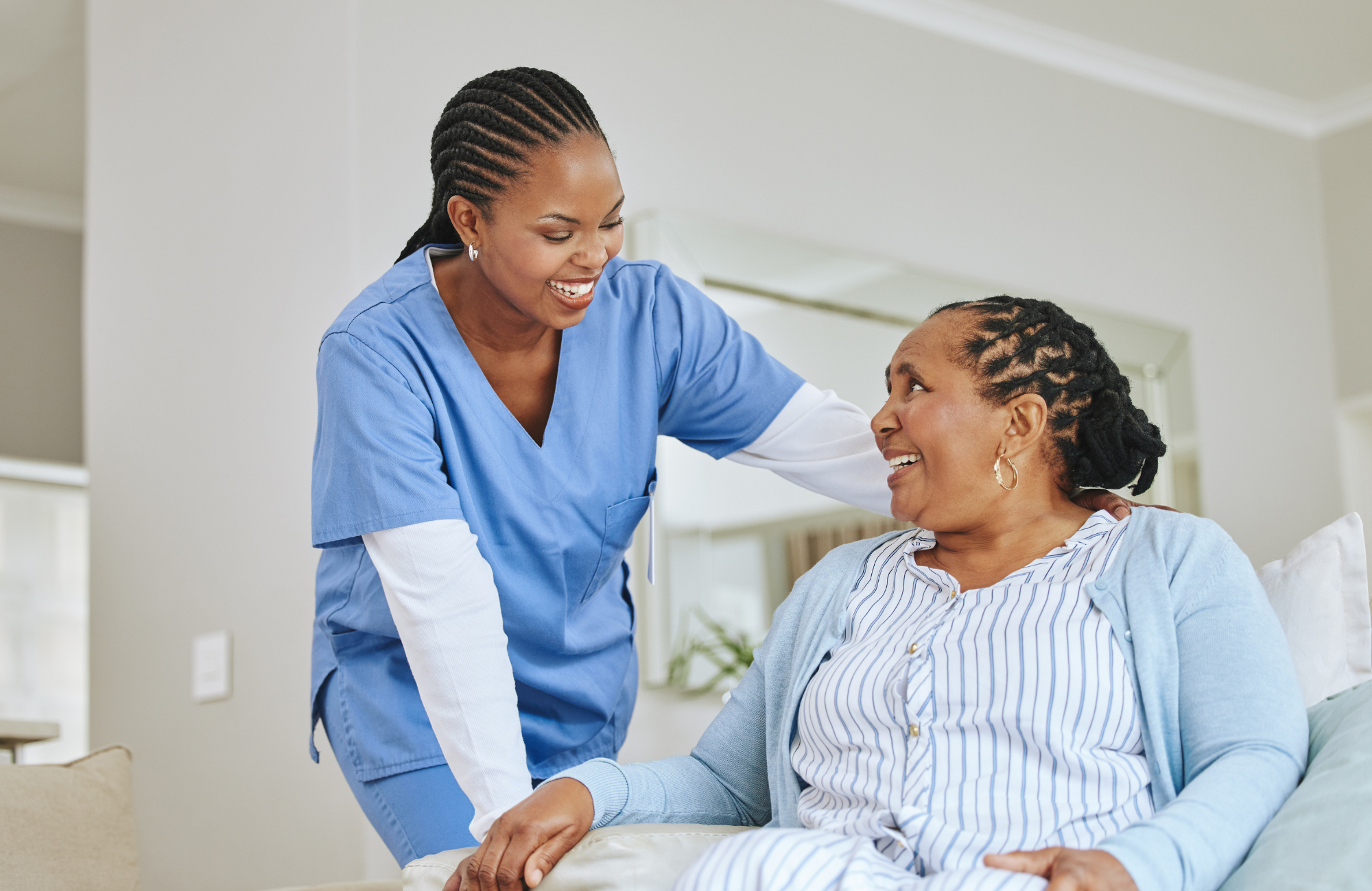 A healthcare professional, wearing blue scrubs, smiles and interacts with a senior woman who is sitting in a hospital bed wearing striped pajamas and a light blue cardigan, in a hospital or healthcare setting.