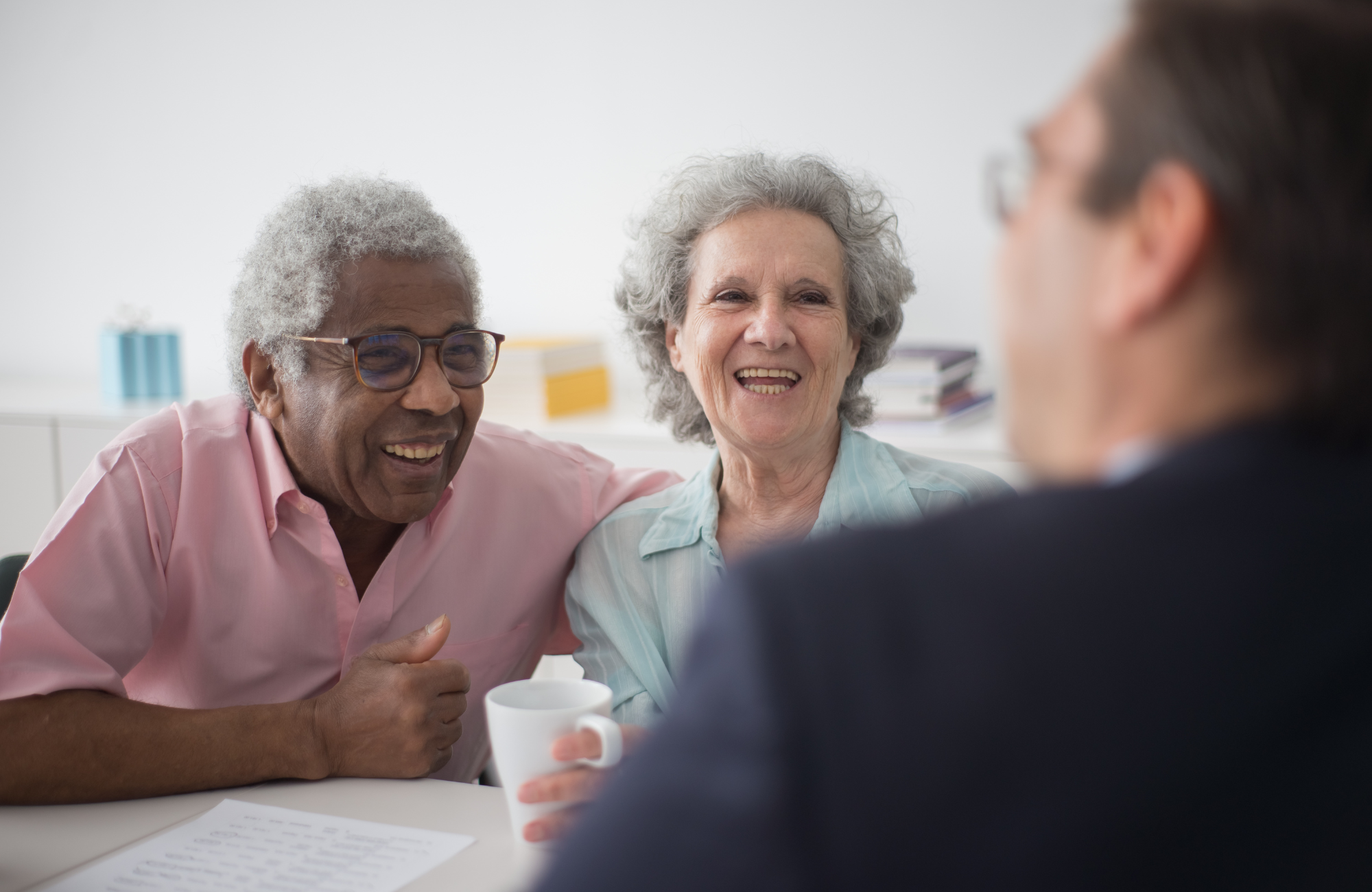 Three older women smiling and engaging in conversation at a table, with a person in the foreground holding a mug.
