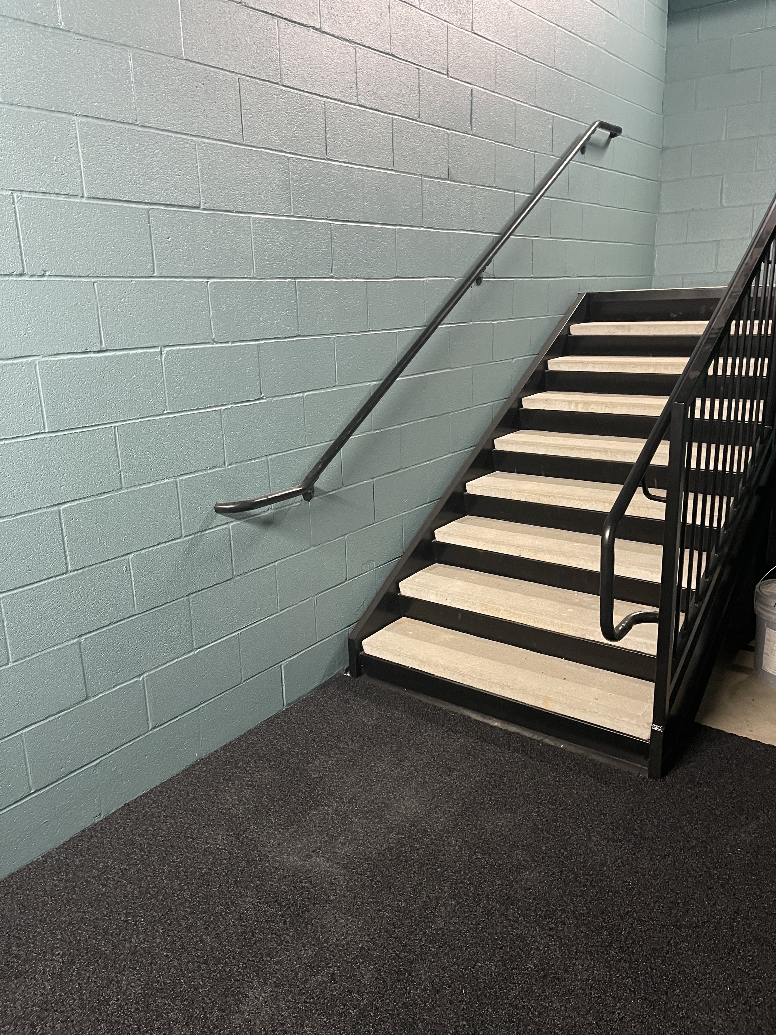 Indoor staircase with black metal railings and beige steps, against a light teal brick wall, with gray carpet flooring at the bottom.
