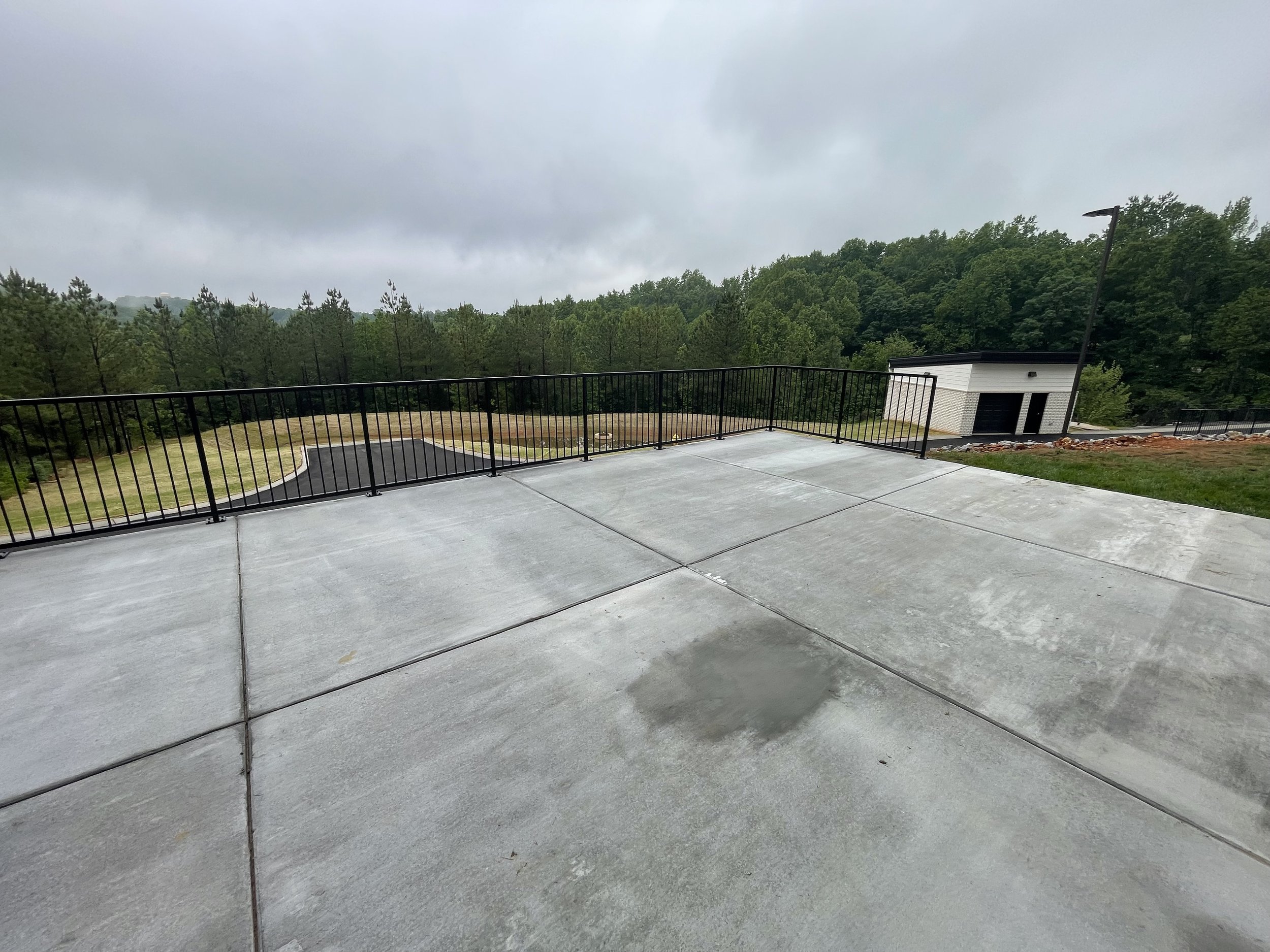 Empty concrete rooftop terrace with black metal railing, overcast sky, and a small building in the background.