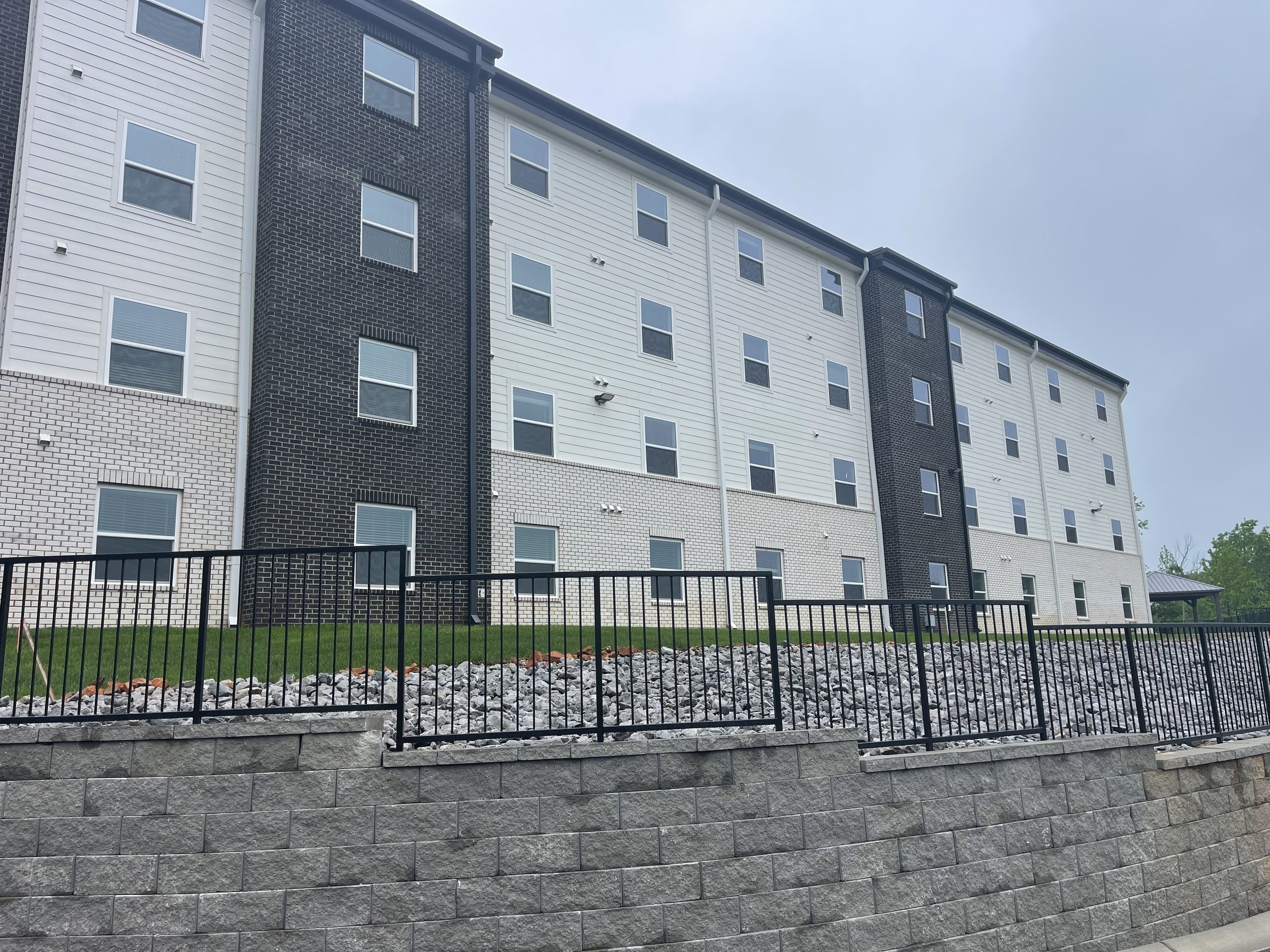 Multi-story residential building with mixed white and black brick exterior, multiple windows, and a black metal fence in front, with a stone retaining wall below.